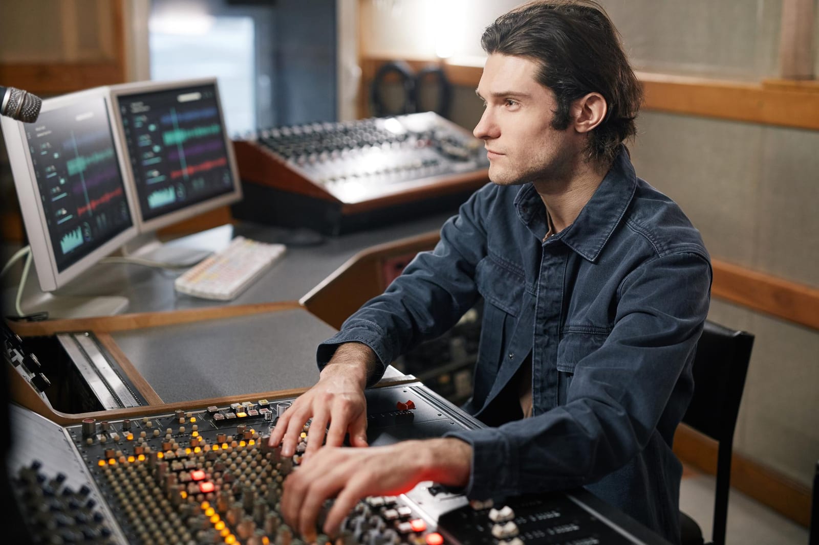Produtor musical trabalha na mesa de som com dois monitores exibindo formas de onda.
